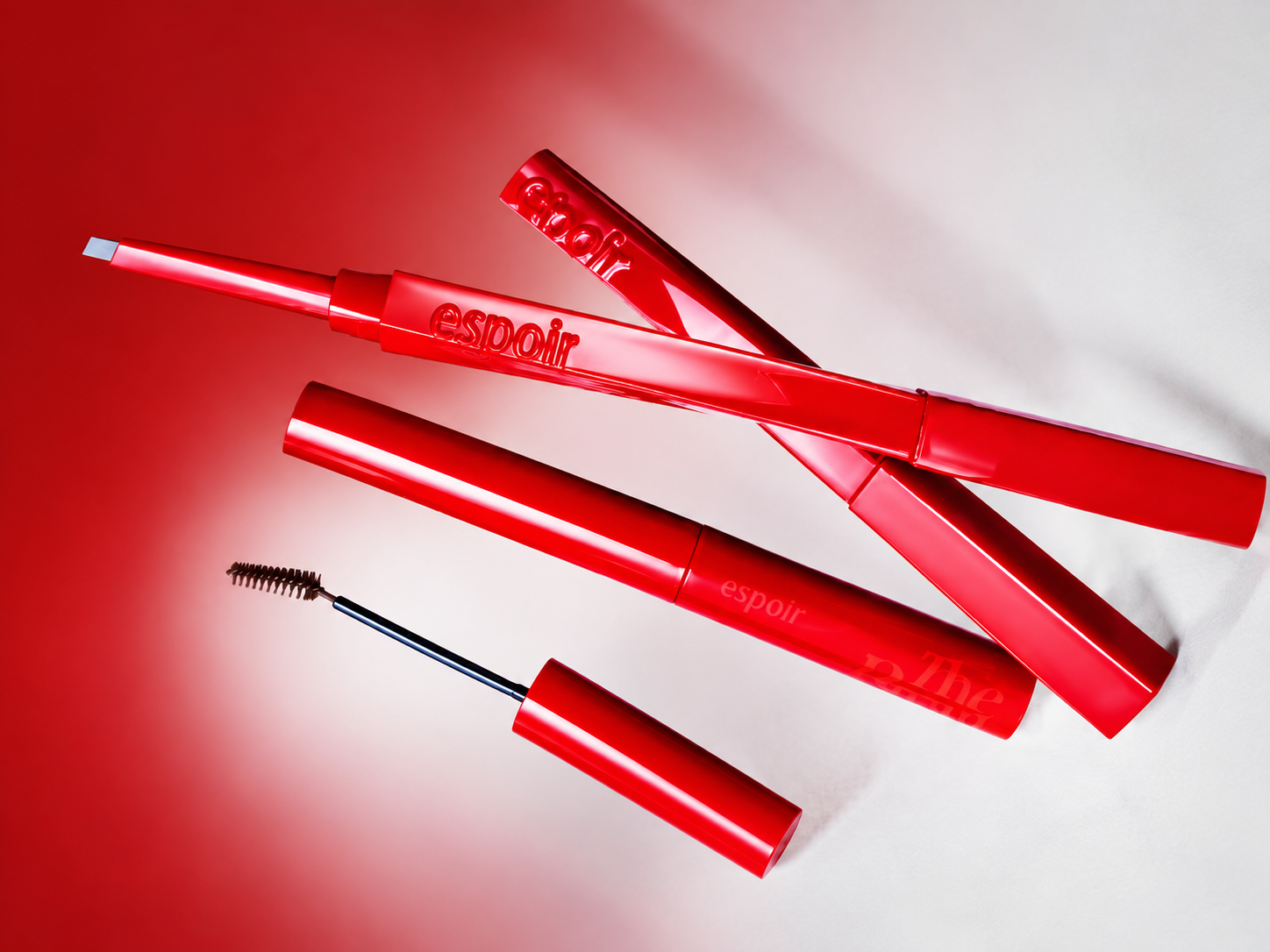 Red espoir makeup pencils and a spoolie brush arranged diagonally on a red and white background.
